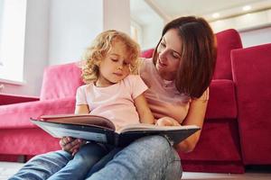 lettura grande libro. giovane madre con sua poco figlia nel casuale Abiti insieme in casa a casa foto