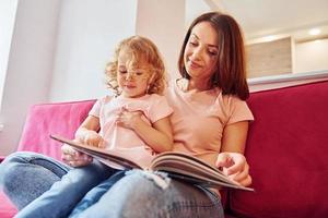 lettura grande libro. giovane madre con sua poco figlia nel casuale Abiti insieme in casa a casa foto