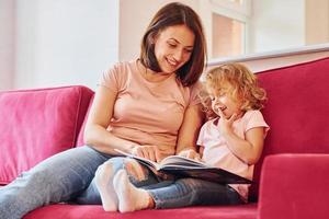 lettura grande libro. giovane madre con sua poco figlia nel casuale Abiti insieme in casa a casa foto