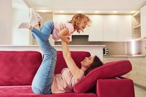 avendo divertimento su rosso divano. giovane madre con sua poco figlia nel casuale Abiti insieme in casa a casa foto