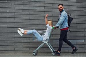 avendo divertimento e equitazione shopping carrello. giovane elegante uomo con donna nel casuale Abiti all'aperto insieme. concezione di amicizia o relazioni foto