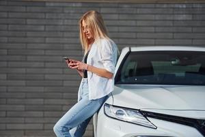 bellissimo bionda nel casuale Abiti con Telefono vicino bianca auto è all'aperto a soleggiato giorno foto