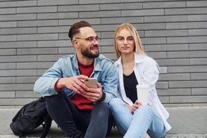 utilizzando Telefono. giovane elegante uomo con donna nel casuale Abiti seduta all'aperto insieme. concezione di amicizia o relazioni foto