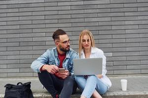 utilizzando il computer portatile. giovane elegante uomo con donna nel casuale Abiti seduta all'aperto insieme. concezione di amicizia o relazioni foto