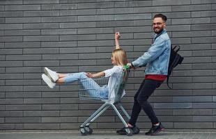 avendo divertimento e equitazione shopping carrello. giovane elegante uomo con donna nel casuale Abiti all'aperto insieme. concezione di amicizia o relazioni foto