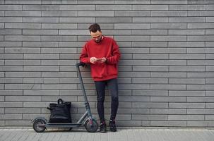 con nero Borsa. bello giovane tipo nel casuale Abiti in piedi con elettrico schooter all'aperto a soleggiato giorno foto