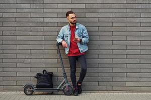 con nero Borsa. bello giovane tipo nel casuale Abiti in piedi con elettrico schooter all'aperto a soleggiato giorno foto