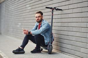 bello giovane tipo nel casuale Abiti seduta su elettrico schooter all'aperto a soleggiato giorno foto