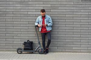 con nero Borsa. bello giovane tipo nel casuale Abiti in piedi con elettrico schooter all'aperto a soleggiato giorno foto