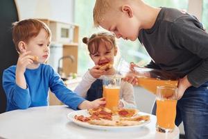 tre bambini seduta in casa di il tavolo e mangiare Pizza con arancia succo insieme foto