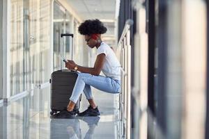 giovane africano americano femmina passanger nel casuale Abiti è nel aeroporto con Bagaglio. utilizzando Telefono foto