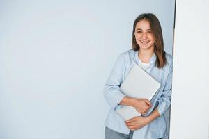 bellissimo giovane donna nel jeans e blu camicia in piedi contro parete con il computer portatile nel mani foto