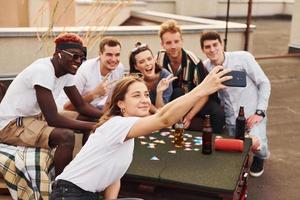 ragazza fare foto quando persone giocando carta gioco. gruppo di giovane persone nel casuale Abiti avere un' festa a tetto insieme a giorno