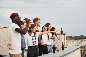 in piedi alto con alcol. nuvoloso cielo. gruppo di giovane persone nel casuale Abiti avere un' festa a tetto insieme a giorno foto