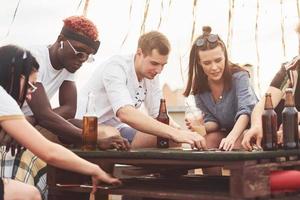 giocando gioco e seduta di il tavolo. gruppo di giovane persone nel casuale Abiti avere un' festa a tetto insieme a giorno foto