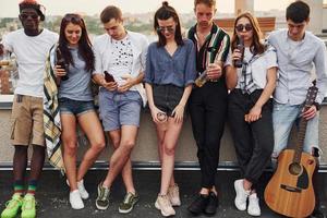 in piedi con telefoni e alcool nel mani. gruppo di giovane persone nel casuale Abiti avere un' festa a tetto insieme a giorno foto