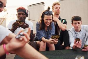 seduta di tavolo e giocando carta gioco. gruppo di giovane persone nel casuale Abiti avere un' festa a tetto insieme a giorno foto