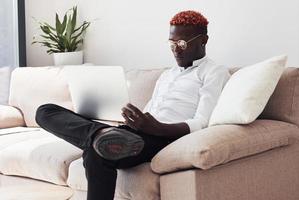 giovane africano americano uomo nel formale Abiti seduta in casa con il computer portatile nel mani foto