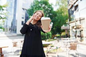 parlando di Telefono e Tenere tazza di bere. contento giovane ragazza nel nero gonna all'aperto nel il città vicino verde alberi e contro attività commerciale edificio foto