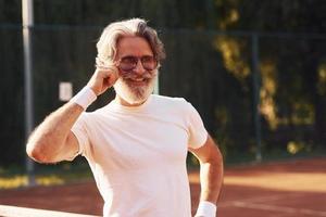 anziano elegante uomo nel occhiali, bianca camicia e nero allegro pantaloncini su tennis Tribunale foto
