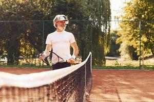 bellissimo luce del sole. anziano elegante uomo nel bianca camicia e nero allegro pantaloncini su tennis Tribunale foto