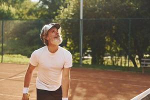 giocando gioco. anziano moderno elegante uomo con racchetta all'aperto su tennis Tribunale a giorno foto