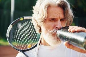 Tenere bottiglia di acqua. anziano moderno elegante uomo con racchetta all'aperto su tennis Tribunale a giorno foto