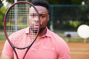 africano americano uomo nel rosa camicia in piedi con tennis racjet su il Tribunale all'aperto foto