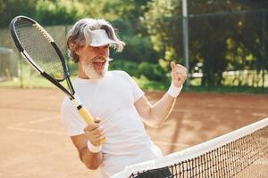 festeggiare vittoria. anziano moderno elegante uomo con racchetta all'aperto su tennis Tribunale a giorno foto