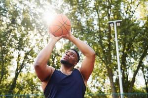 bellissimo verde alberi su sfondo. africano americano uomo giochi pallacanestro su il Tribunale all'aperto foto