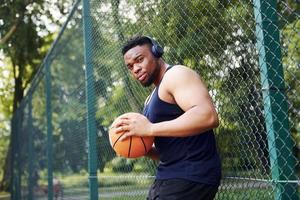 africano americano uomo con senza fili cuffie prende un' rompere e pendente su il metallo maglia con palla su il Tribunale all'aperto foto