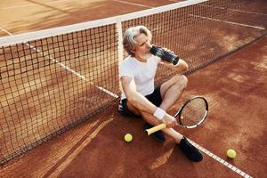 potabile acqua e seduta su il terra. anziano moderno elegante uomo con racchetta all'aperto su tennis Tribunale a giorno foto