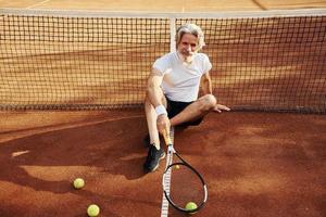 si siede su il terra e prende rompere. anziano moderno elegante uomo con racchetta all'aperto su tennis Tribunale a giorno foto