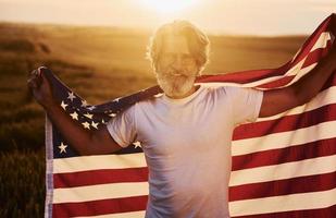 concezione di libertà. Tenere Stati Uniti d'America bandiera. anziano elegante uomo con grigio capelli e barba su il agricolo campo con raccogliere foto