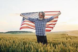 Tenere Stati Uniti d'America bandiera nel mani. patriottico anziano elegante uomo con grigio capelli e barba su il agricolo campo foto