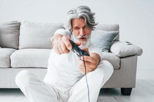 giochi video gioco di utilizzando controllore. anziano elegante moderno uomo con grigio capelli e barba in casa foto