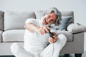 giochi video gioco di utilizzando controllore. anziano elegante moderno uomo con grigio capelli e barba in casa foto