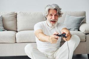 giochi video gioco di utilizzando controllore. anziano elegante moderno uomo con grigio capelli e barba in casa foto