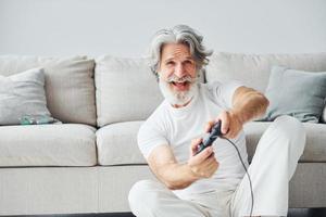 giochi video gioco di utilizzando controllore. anziano elegante moderno uomo con grigio capelli e barba in casa foto