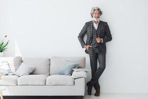 nel elegante Abiti. in piedi contro bianca parete. anziano elegante moderno uomo con grigio capelli e barba in casa foto