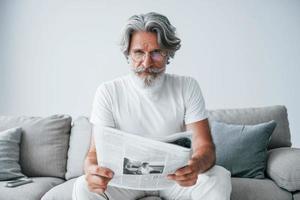 si siede su il divano. anziano elegante moderno uomo con grigio capelli e barba in casa foto