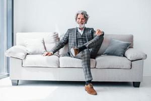 si siede su confortevole divano nel formale Abiti. anziano elegante moderno uomo con grigio capelli e barba in casa foto