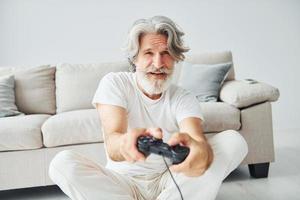 giochi video gioco di utilizzando controllore. anziano elegante moderno uomo con grigio capelli e barba in casa foto