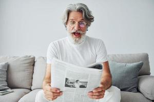 si siede su il divano. anziano elegante moderno uomo con grigio capelli e barba in casa foto