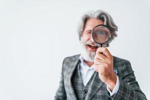 avendo divertimento con ingrandimento bicchiere. anziano elegante moderno uomo con grigio capelli e barba in casa foto