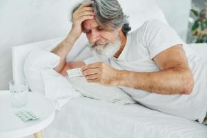 malato anziano elegante moderno uomo con grigio capelli e barba in casa dire bugie su il letto e si sente cattivo foto