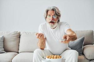 Guardando tv mostrare e mangia Popcorn. anziano elegante moderno uomo con grigio capelli e barba in casa foto