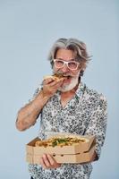 mangia Pizza quando in piedi contro parete. anziano elegante moderno uomo con grigio capelli e barba in casa foto