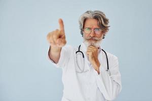 mostrando qualcosa di dito. medico lavoratore nel cappotto. anziano elegante moderno uomo con grigio capelli e barba in casa foto