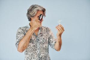 avendo divertimento con ingrandimento bicchiere. anziano elegante moderno uomo con grigio capelli e barba in casa foto